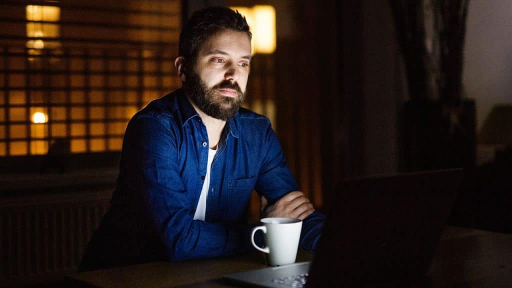 A bearded man in a blue shirt works on a laptop at night, a mug nearby.