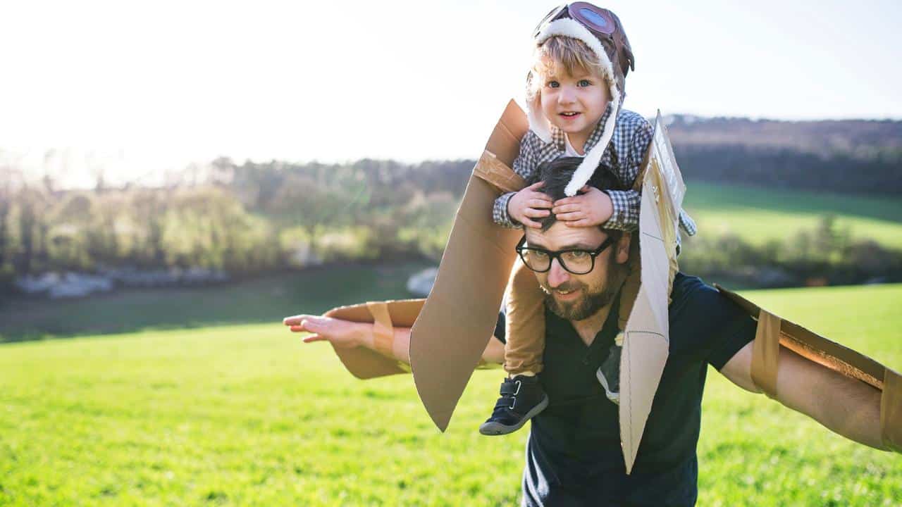 A man giving a piggyback ride to a child wearing cardboard wings in a field.
