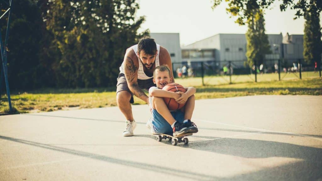 A man pushing a boy sitting on a skateboard holding a basketball.