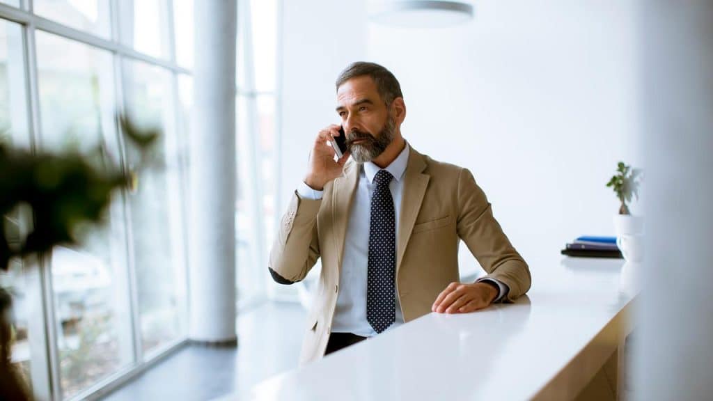 A bearded man in a tan suit talking on a smartphone at a reception counter.