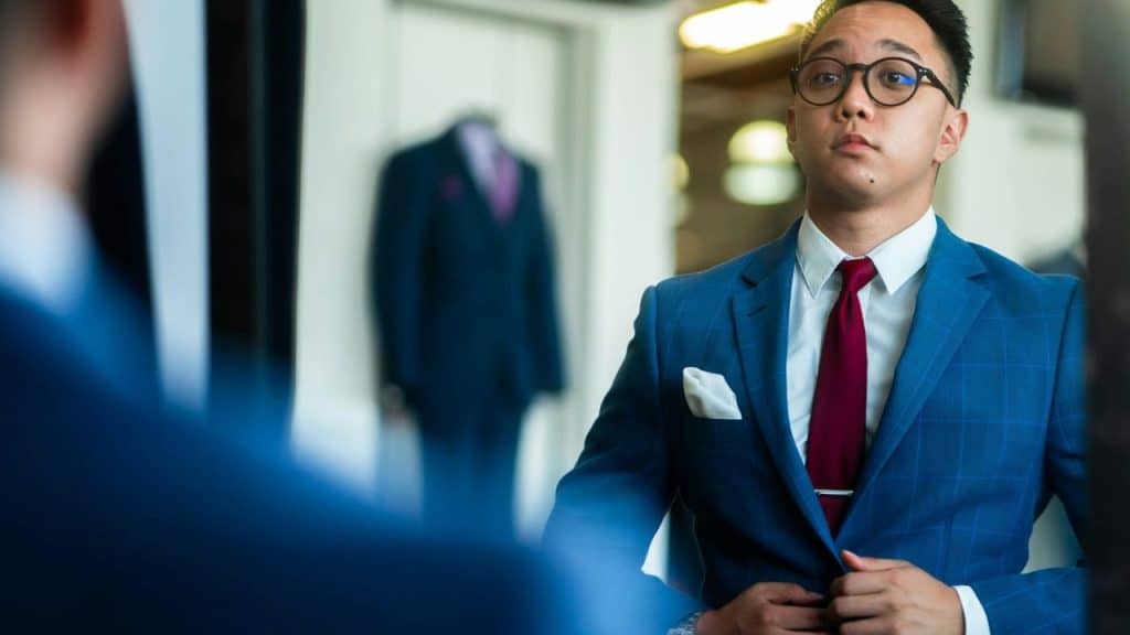 A man in glasses and a blue suit adjusts his jacket, looking at a mirror.