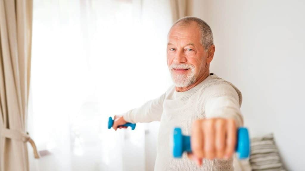 A smiling older man with a beard and a light-colored sweater exercises with blue dumbbells.