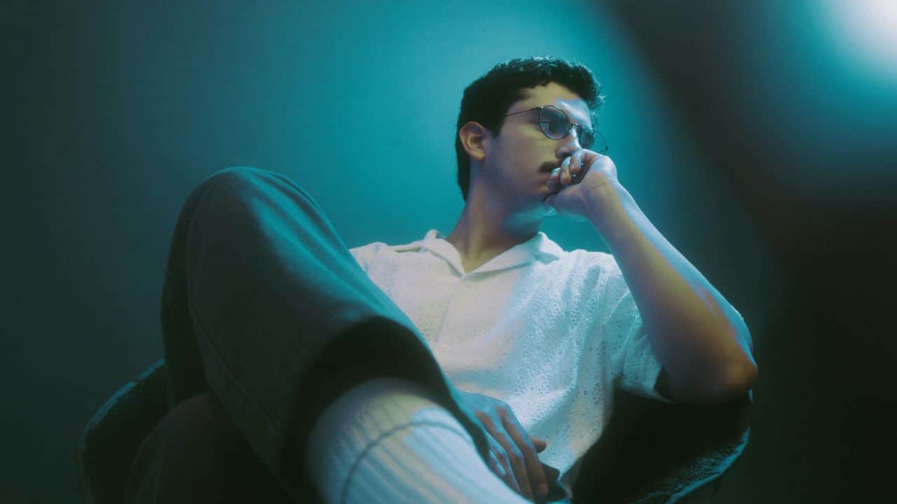 A person in a white collared shirt and glasses sits, looking thoughtfully to their right.