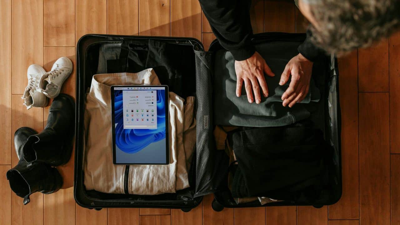 A person packs a black suitcase with clothes and a tablet on a wooden floor, with shoes nearby.