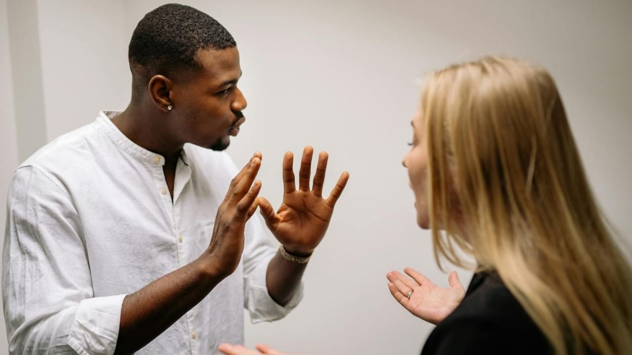 A Black man and a white woman are engaged in a heated argument, gesturing with their hands.