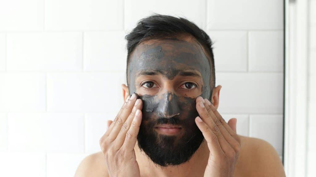 A man applying a black facial mask in a tiled bathroom.