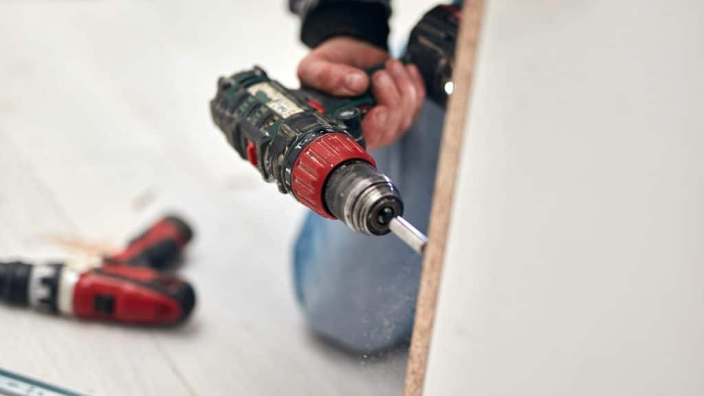 A red-and-black cordless drill driving a screw into a wooden panel.