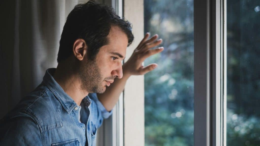 A sad man in a denim shirt stares out of a window with his hand on the pane.