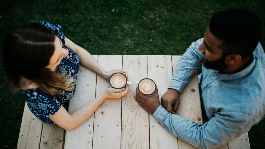 An overhead view shows a man and woman holding coffee cups at a wooden table.