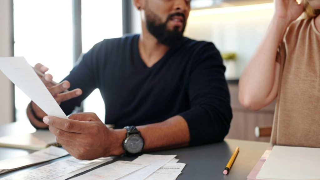 A person with a beard and watch holds papers, discussing them with another person.