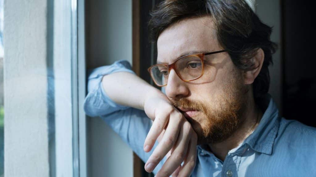 A man with glasses and a beard, in a blue shirt, leans by a window looking away.