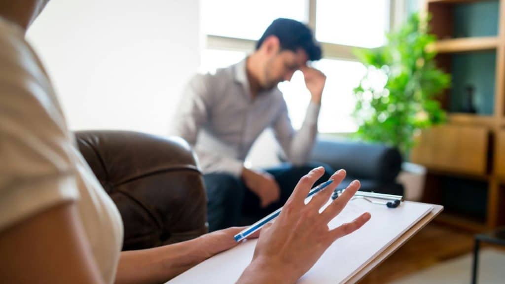 A person holds a clipboard while a man sits in the background, looking distressed.