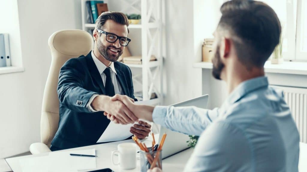 A smiling man in a suit and glasses shakes hands with another man across a desk.