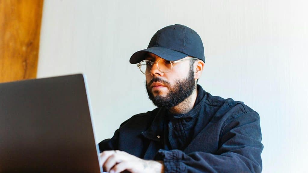 A bearded man in a cap working on a laptop.