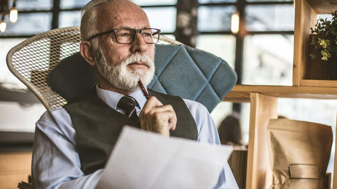 A contemplative, bearded man in glasses and a vest holds a pen to his chin.