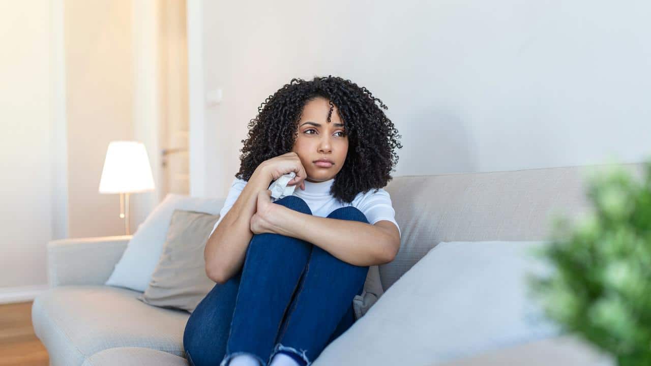 A woman sitting on a couch holding a tissue and looking distressed.