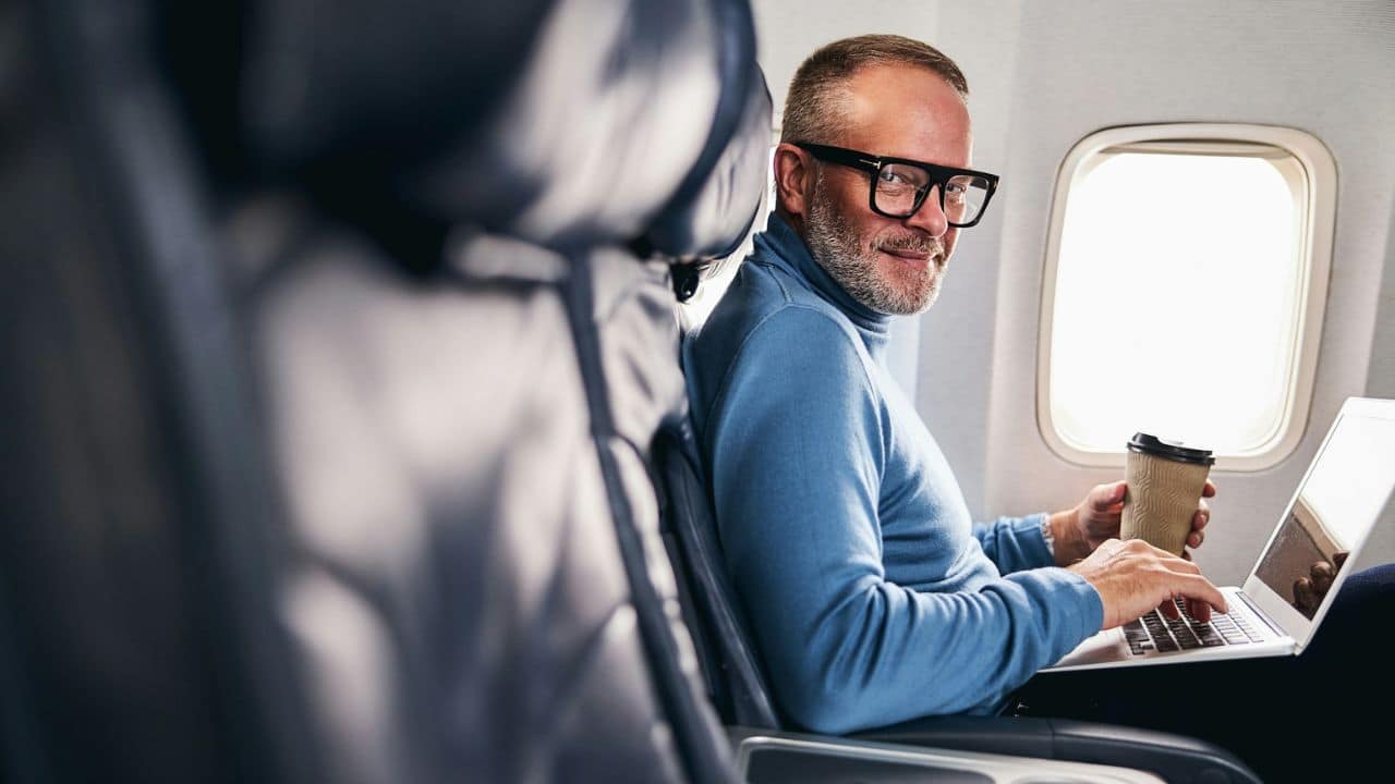 A smiling man with glasses and a beard works on a laptop on an airplane.