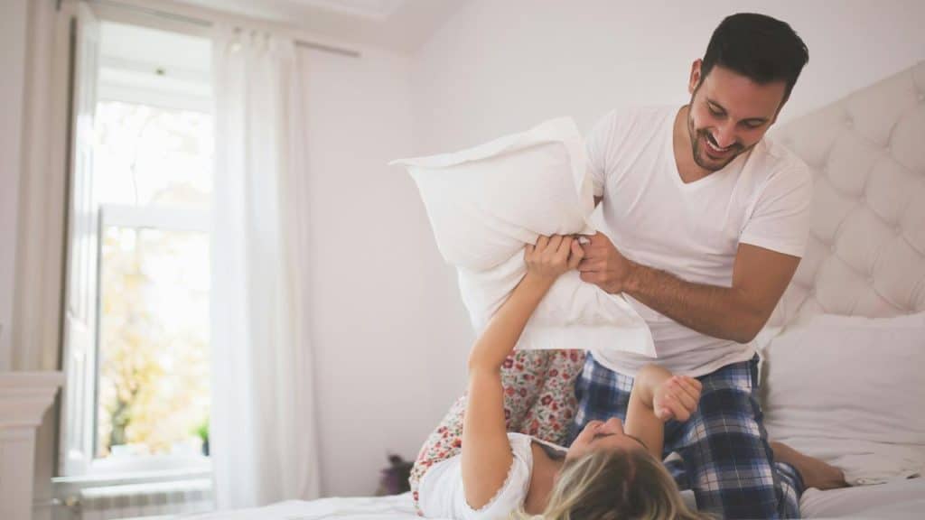A man and woman laughing as they have a pillow fight on a bed.