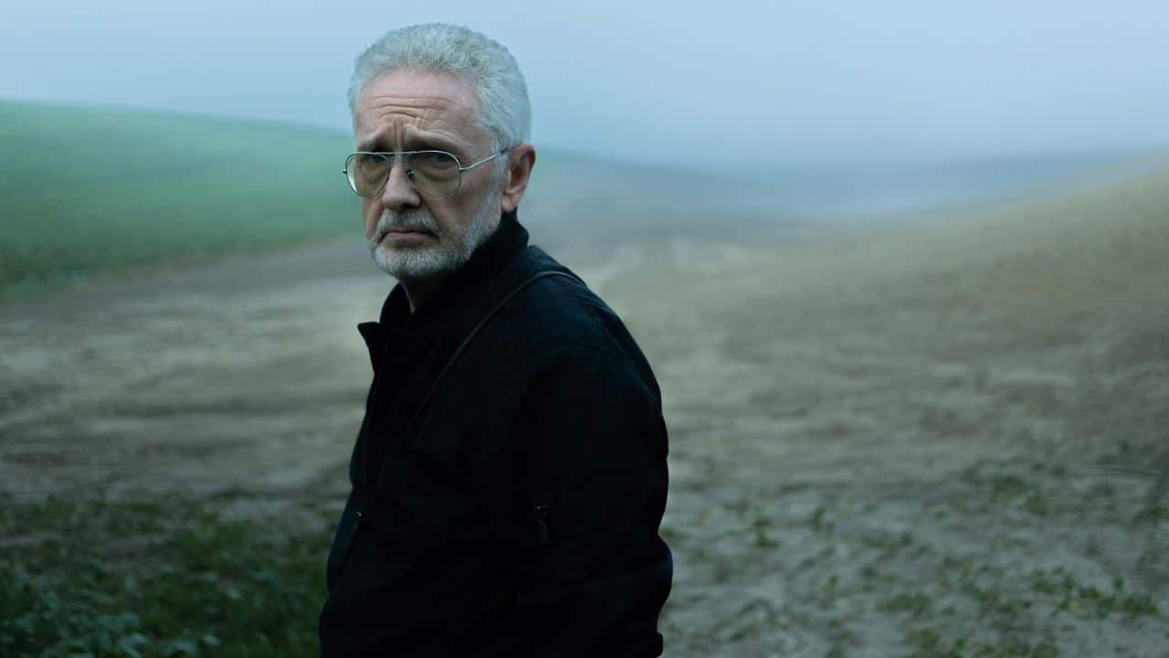 A senior man with glasses and a beard stands in a misty, rural field.