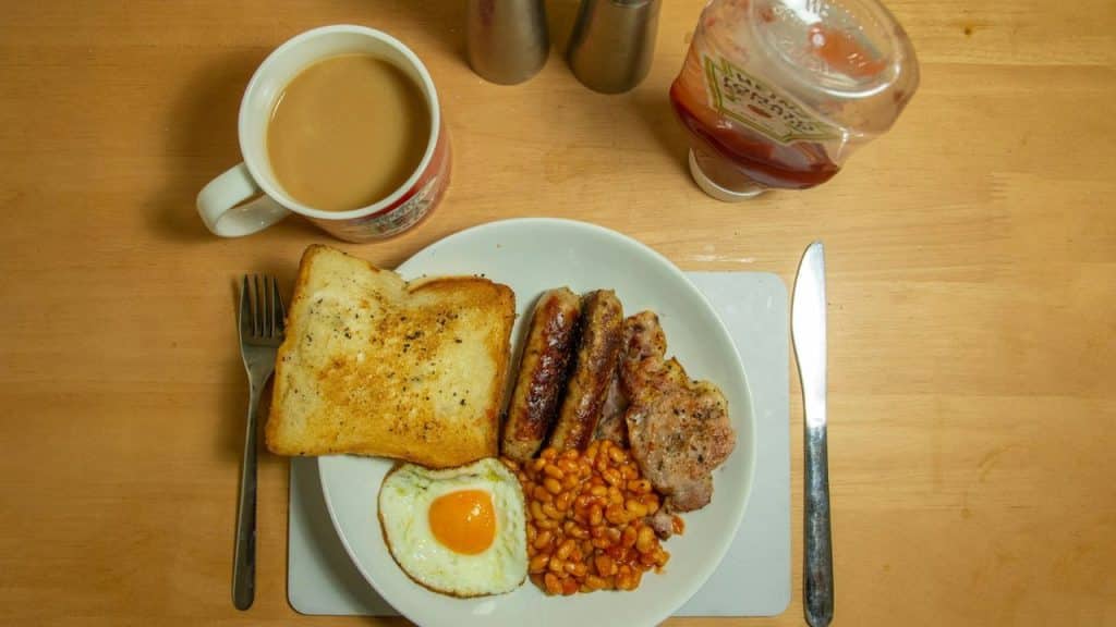 A full English breakfast with toast, sausage, egg, beans, and coffee on a wooden table.