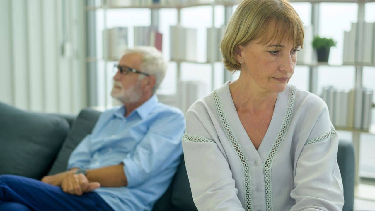 A distressed woman sits in the foreground, with a man blurred in the background.