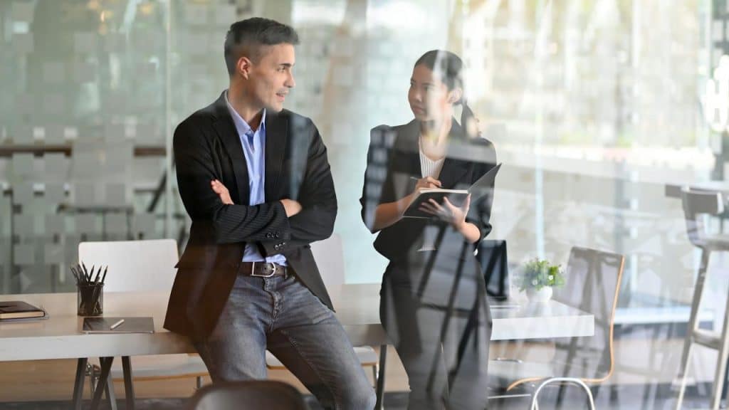 A man leans on a desk while a woman holds a notebook in a modern office.