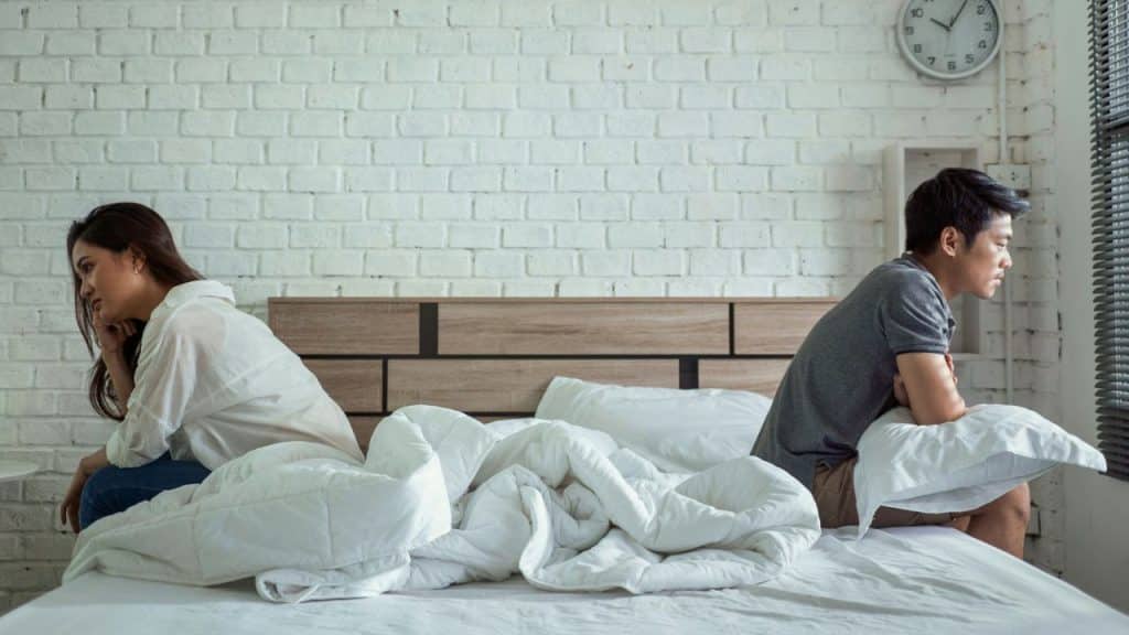 A man and a woman sit on opposite sides of a bed, looking away from each other.