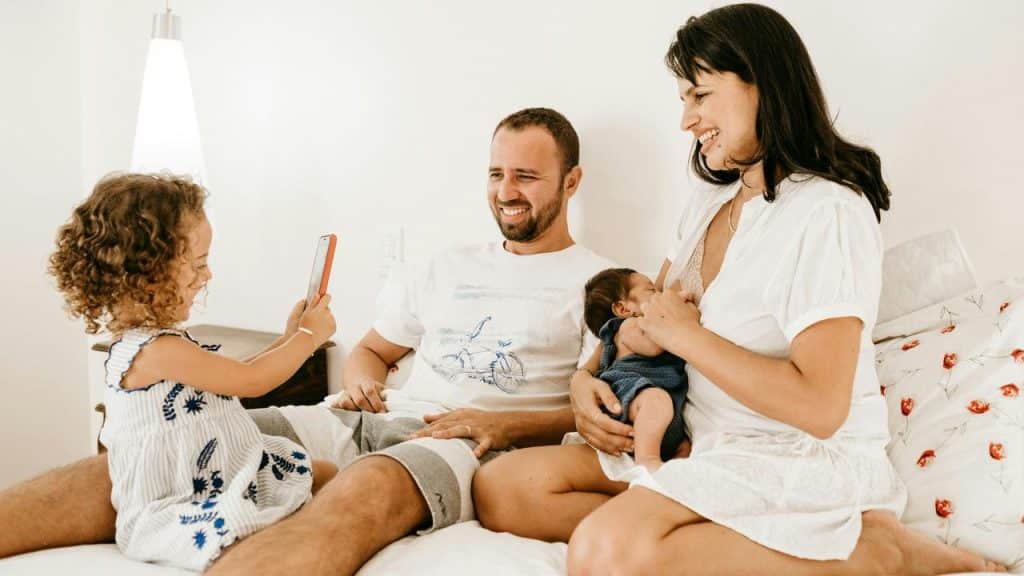 A family on a bed with a mother breastfeeding and a child taking a photo.