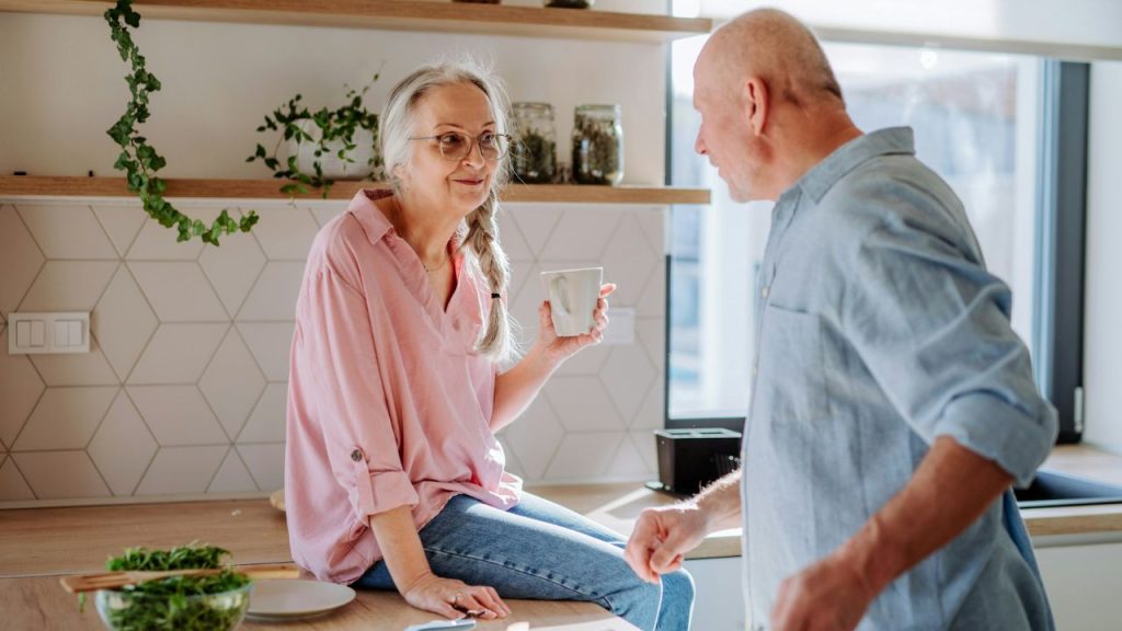 A happy elderly couple converses in a bright kitchen, holding mugs.