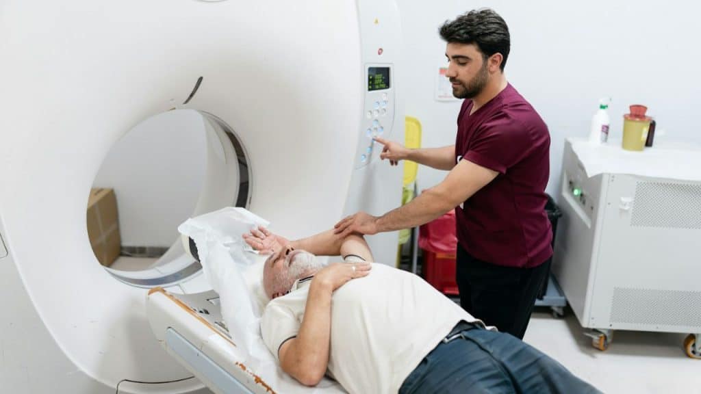A male technician helps a male patient lie on a CT scanner bed.