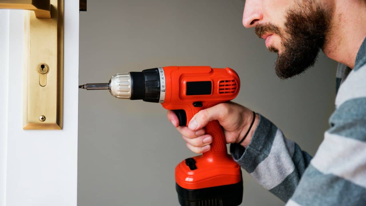 A man with a beard uses a red cordless drill to tighten a screw on a gold door handle.