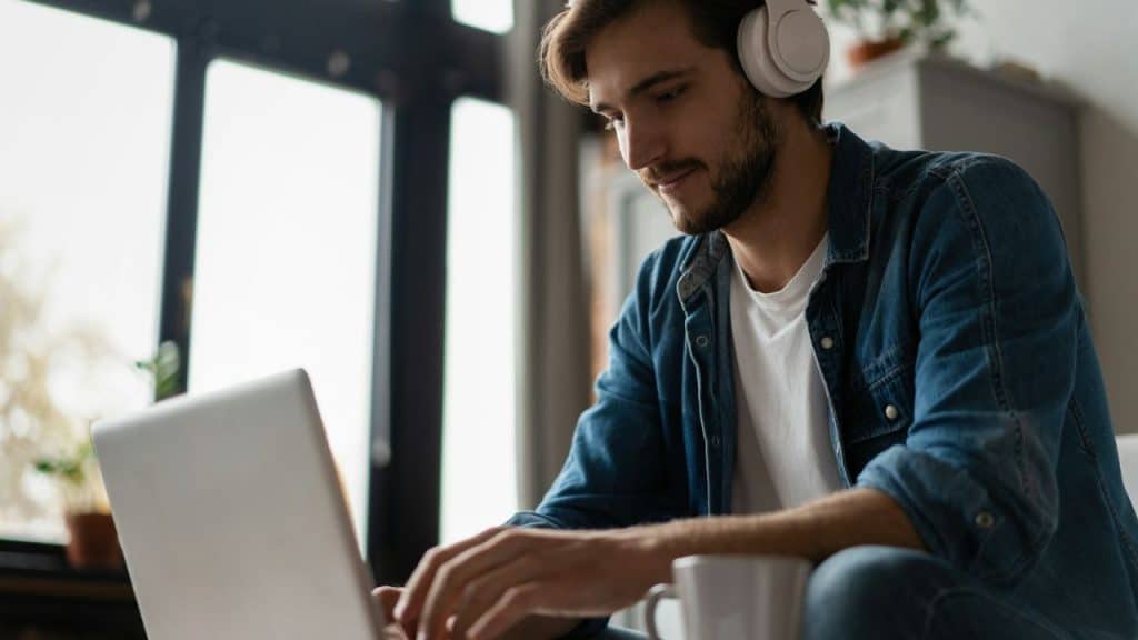 A man wearing headphones uses a laptop, with a mug nearby.