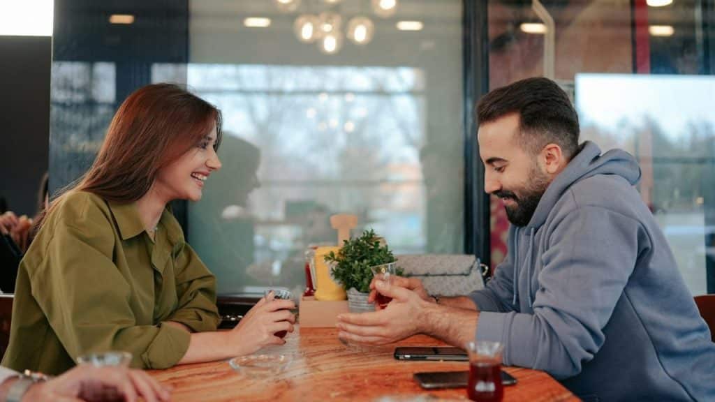 A smiling woman looks at a bearded man holding a glass, both at a table.