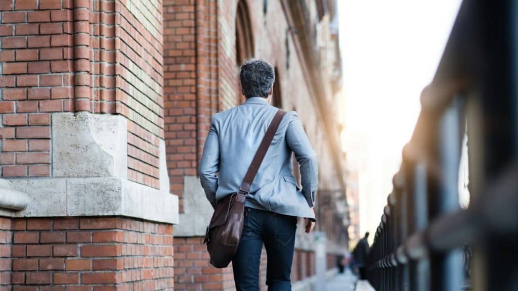 A man with a satchel over his shoulder walks away from the camera down a sunny street next to a brick building.
