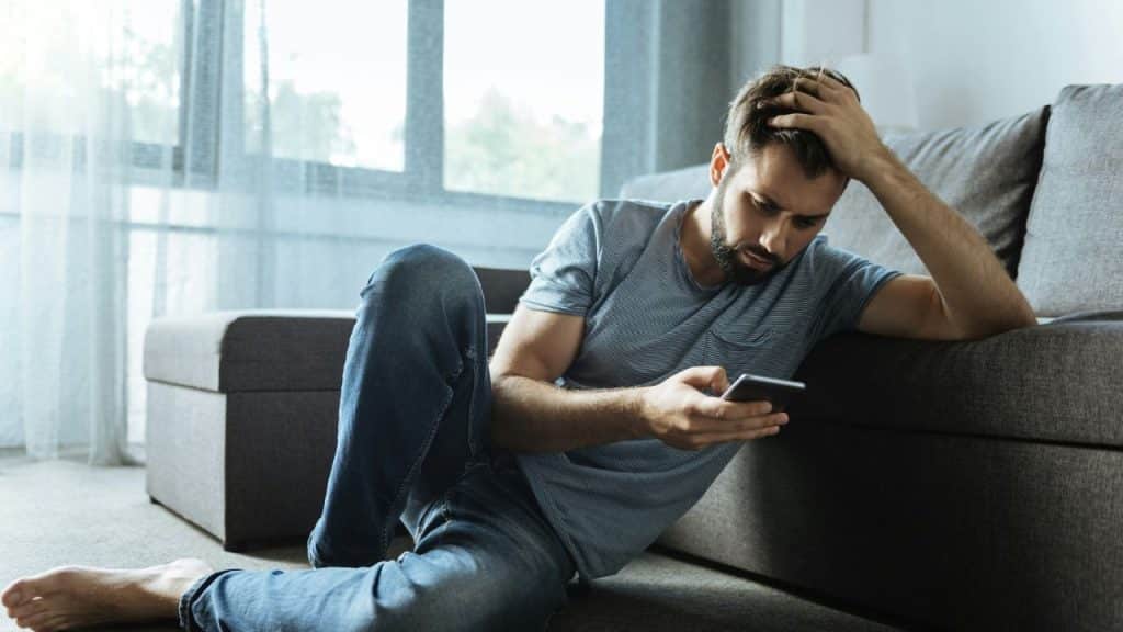 A pensive man sits on the floor, leaning on a sofa, looking at his phone.