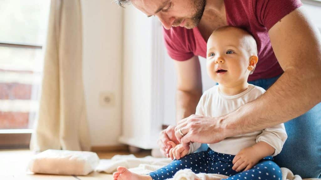 A father helping his baby sit on a blanket.
