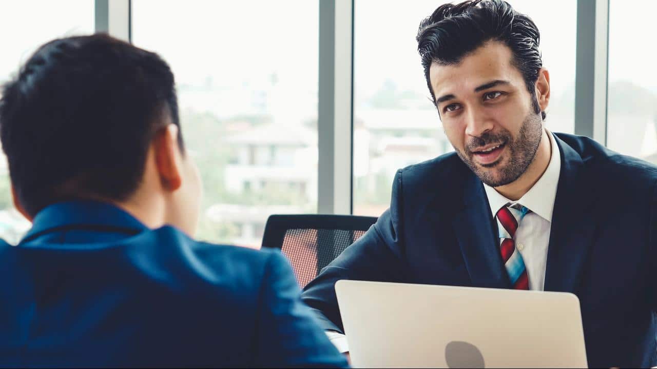 A suited man speaking to a colleague over a laptop.