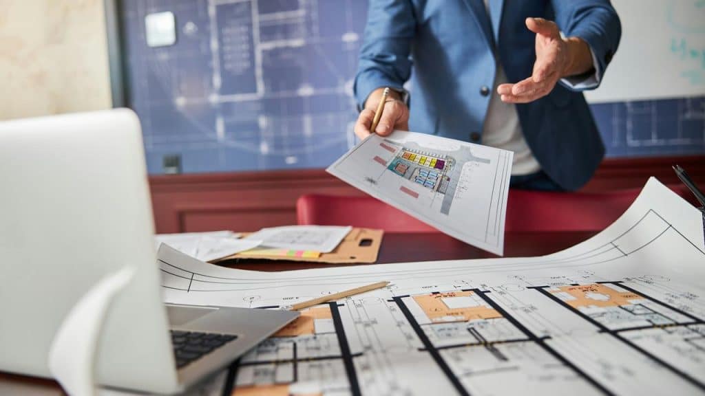 An person presenting a colored floor plan over a desk covered in blueprints and a laptop.
