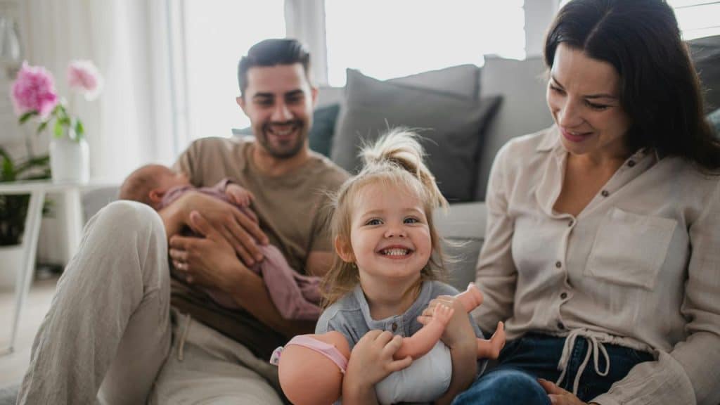 A toddler holding a doll with smiling parents and baby on a couch.