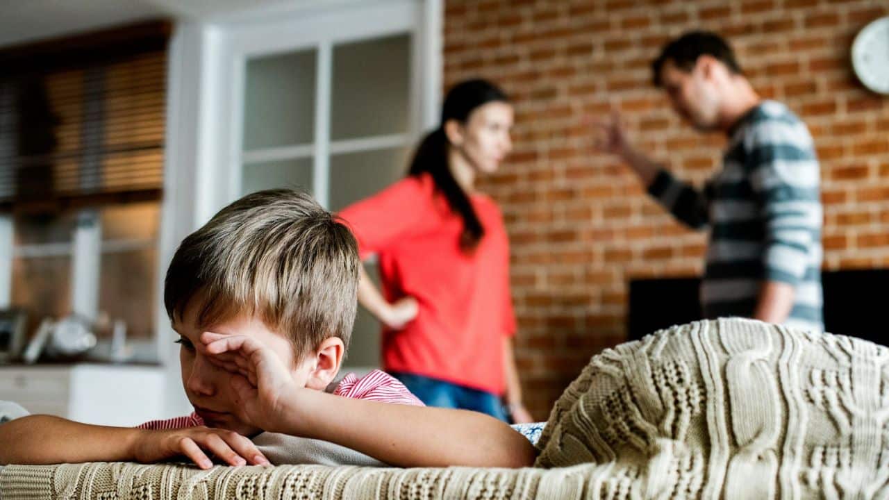 A boy with a worried expression lies on a couch, with arguing adults in the background.