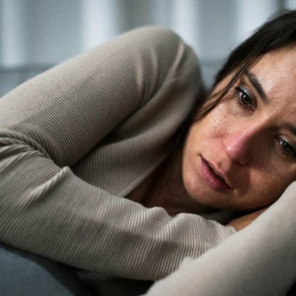 A distressed woman with dark hair and a beige top lies on a grey couch, looking thoughtful.