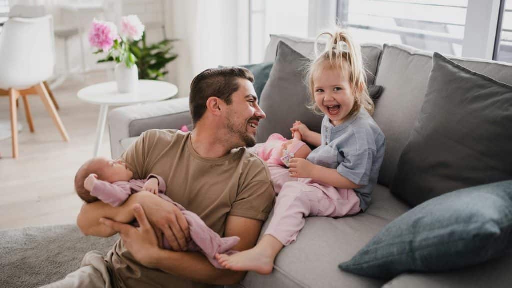 A smiling father holding a newborn as his giggling toddler sits beside him on a couch.