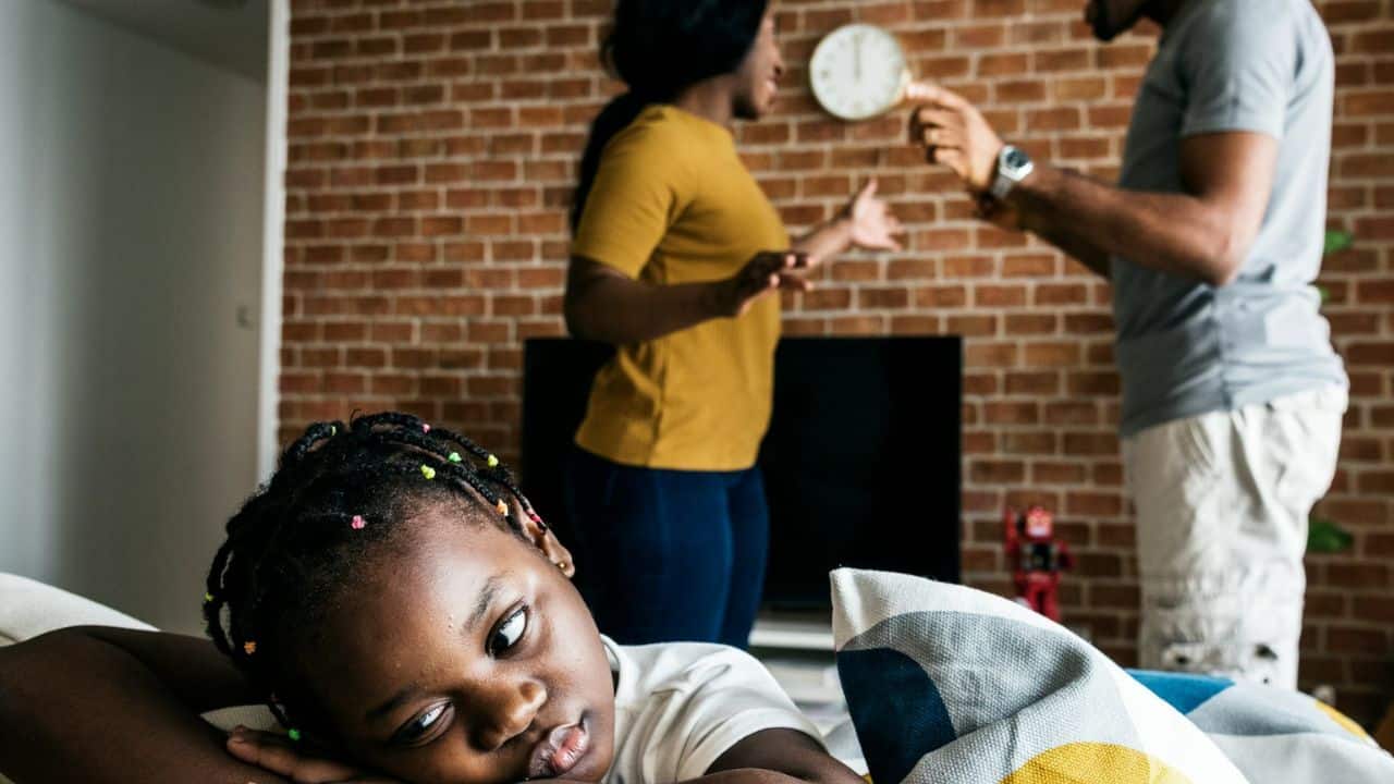 A young child lies in the foreground, looking distressed, while two adults argue in the background.