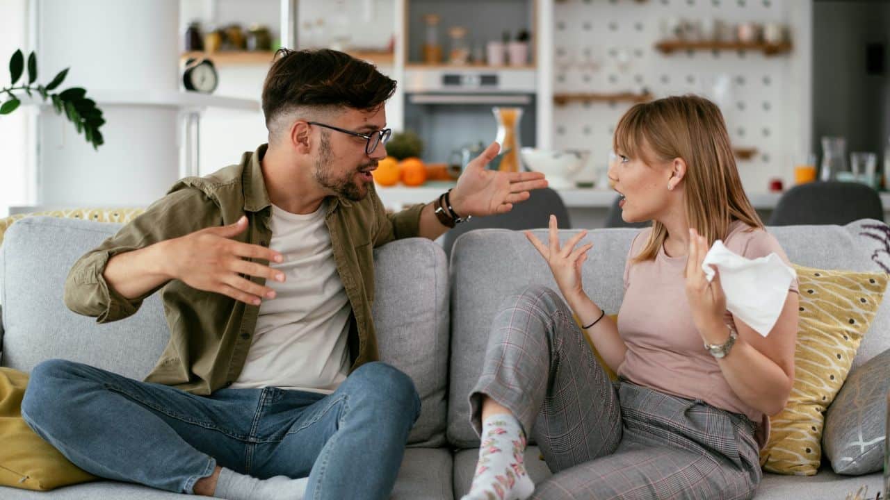 A man and a woman are arguing on a sofa, gesturing expressively.