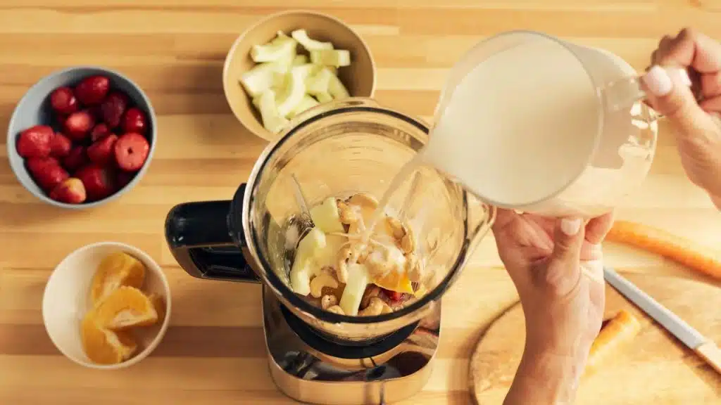 A blender filled with fruit and cashews as milk is poured in.