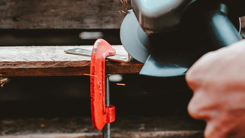 A red C‑clamp holding a wooden plank while an angle grinder cuts metal, sending sparks.