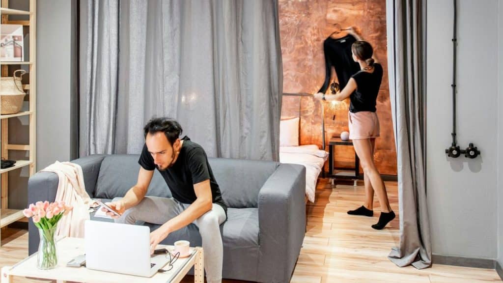 A man works on a laptop on a sofa while a woman gets dressed in the background.