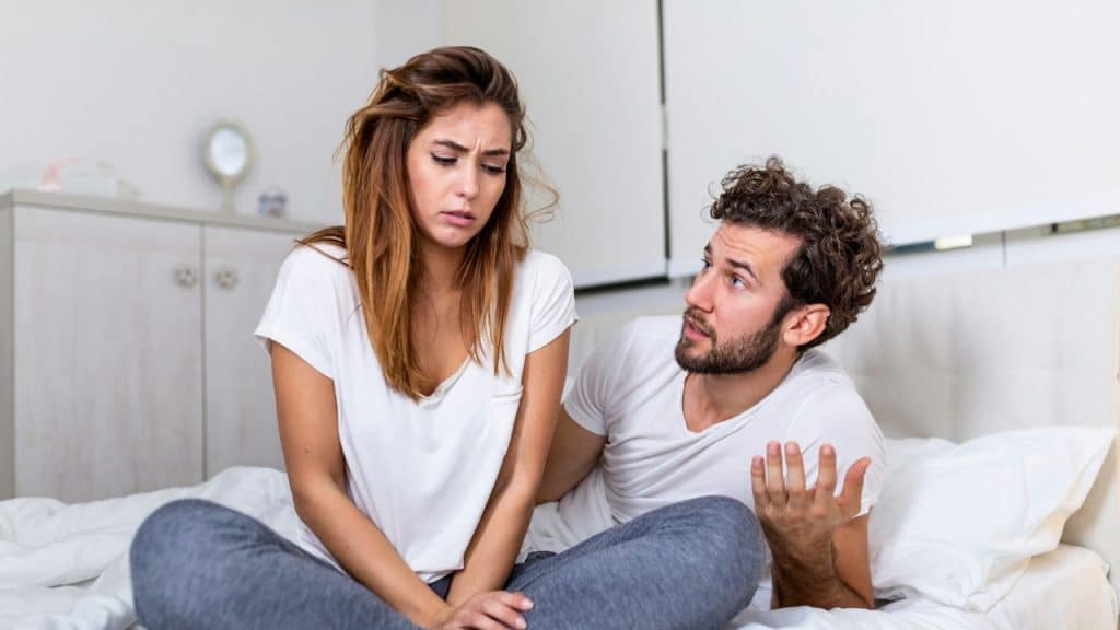 A man sitting in bed gestures at a sad-looking woman next to him.