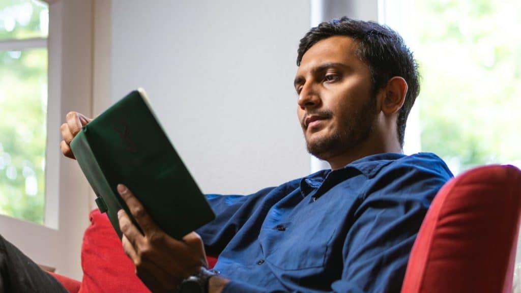 A man with a beard sits in a red chair, reading a green book.