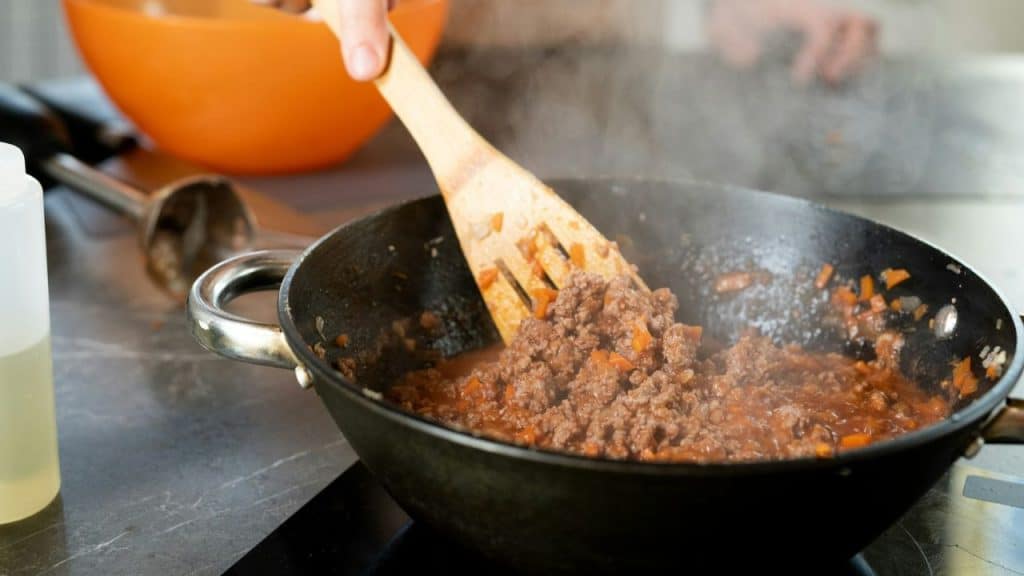 A wooden spoon stirs simmering ground meat and vegetables in a heavy-duty pan.